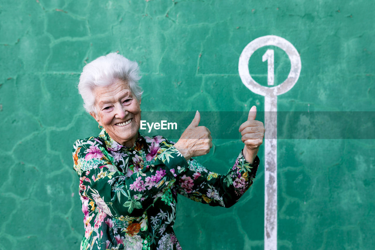 Happy aged gray haired female in colorful blouse standing against green wall and pointing at number 1 sign on parking lot