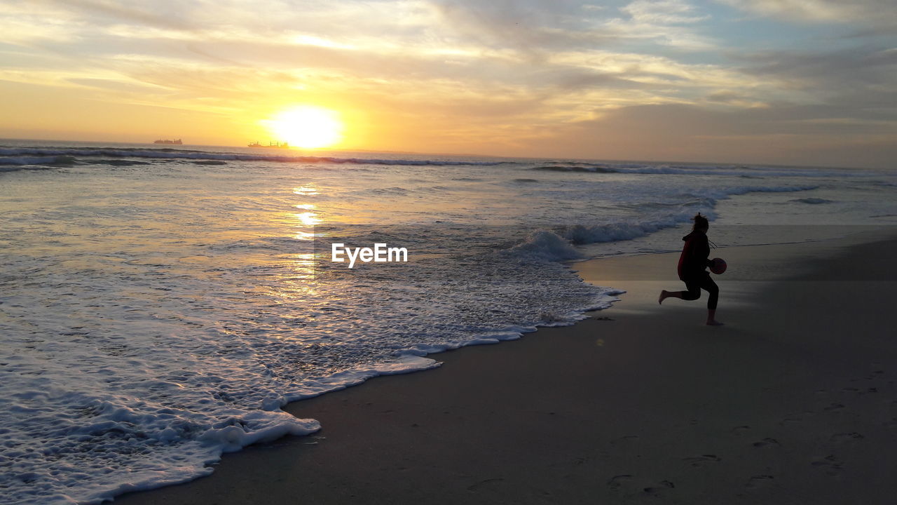 Girl playing at beach against sky during sunset