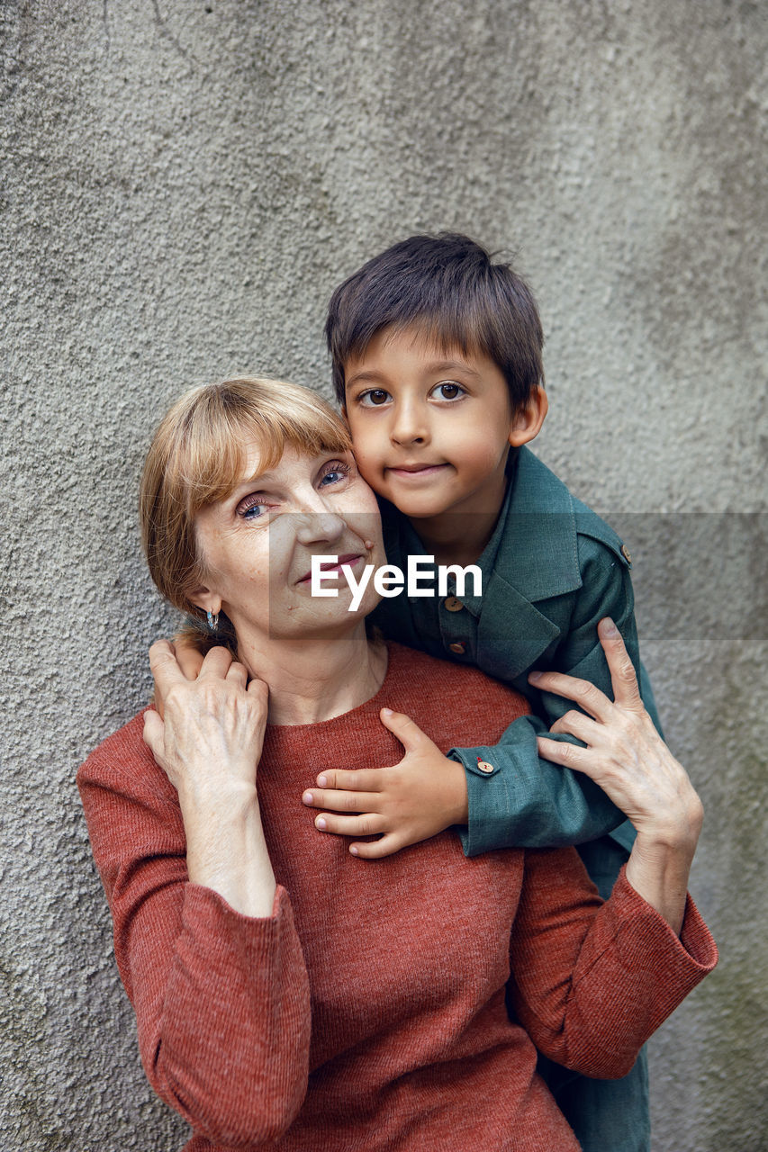 Child boy hugs his grandmother an elderly woman on the street against a gray wall