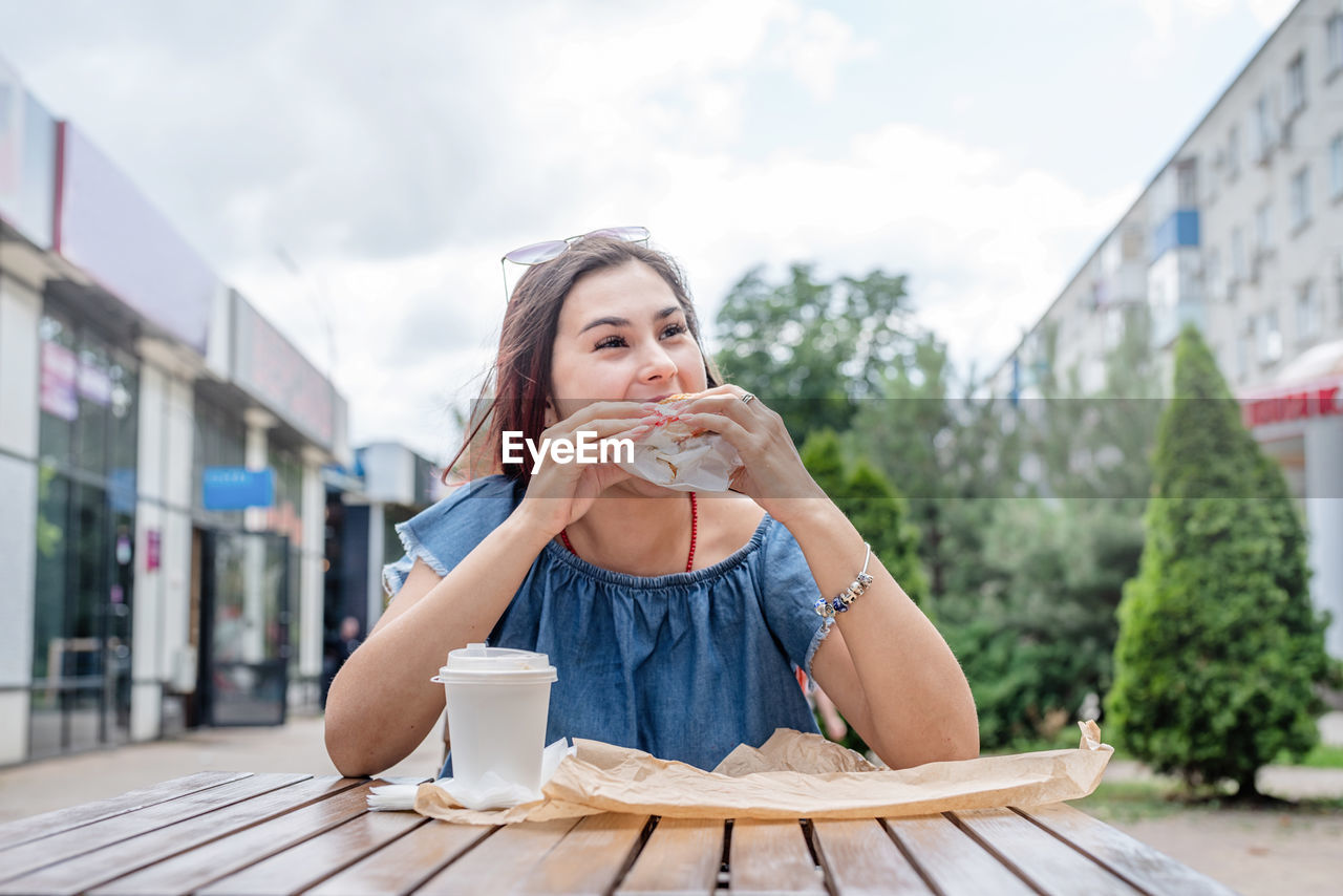 Young woman drinking coffee eating burger while sitting in street cafe