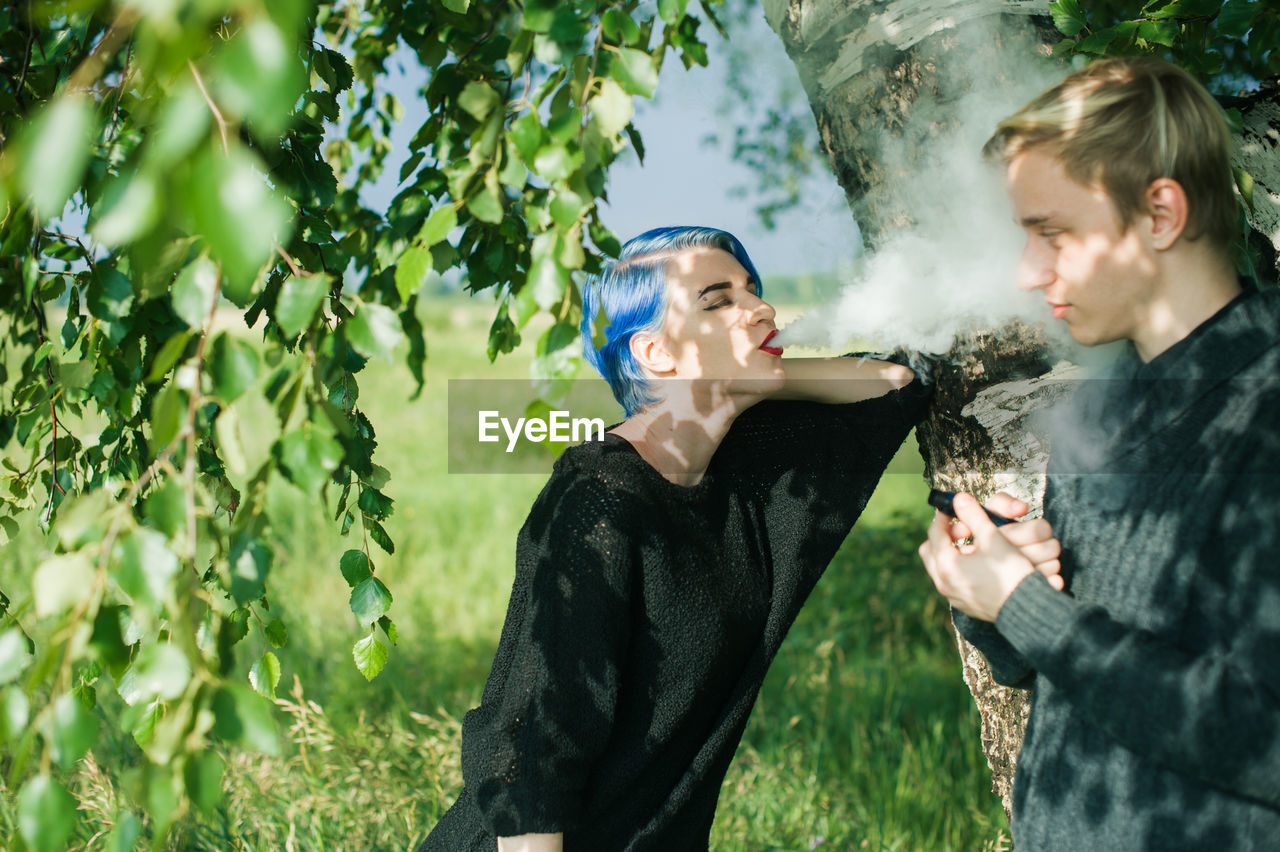 Young woman exhaling smoke on friend by tree
