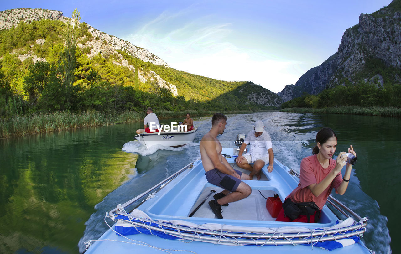 PEOPLE SITTING ON BOAT IN LAKE BY MOUNTAINS AGAINST SKY