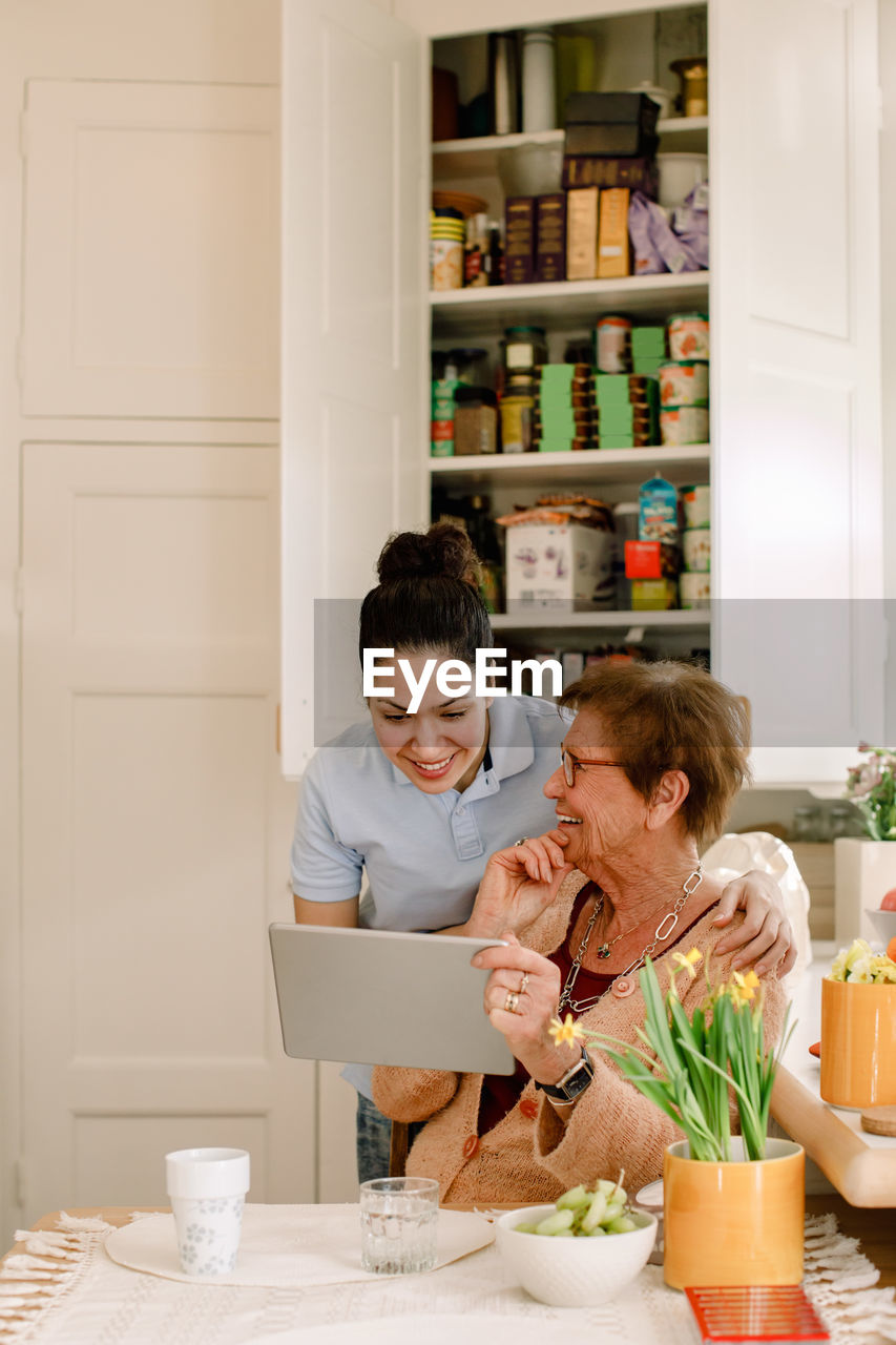 Smiling elderly woman sharing digital tablet with young female caregiver working in kitchen at nursing home