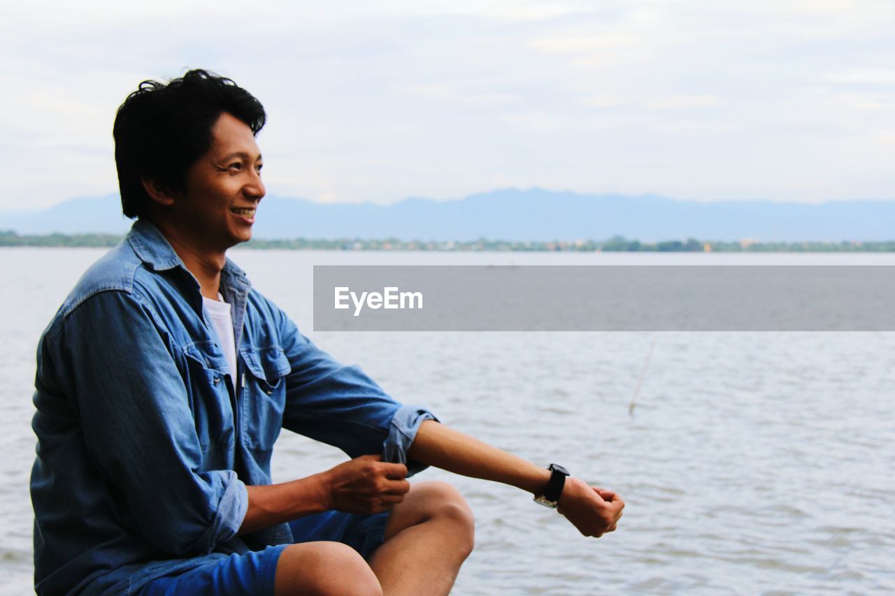 Smiling man sitting by sea against sky