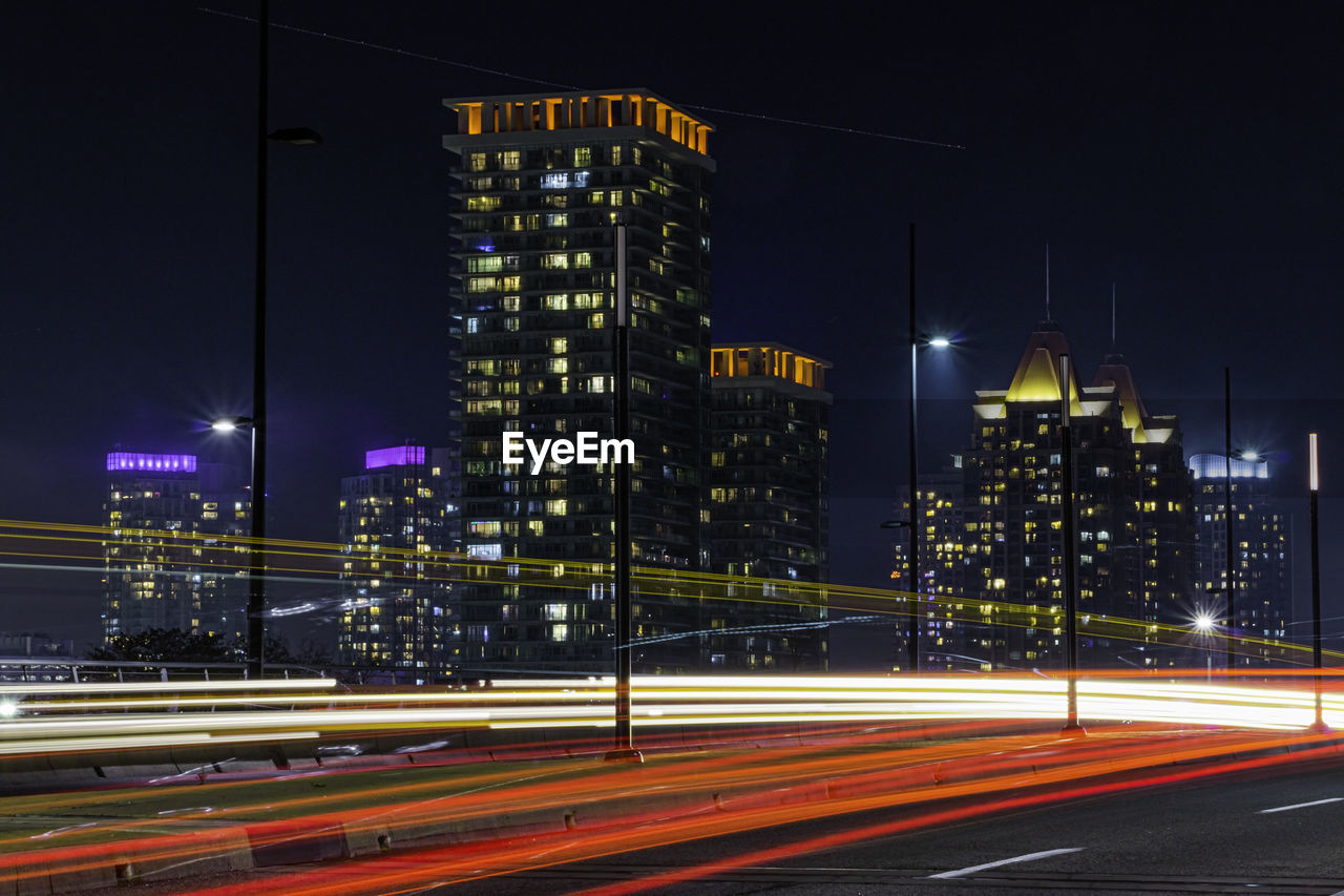 Light trails on road by illuminated buildings in city at night