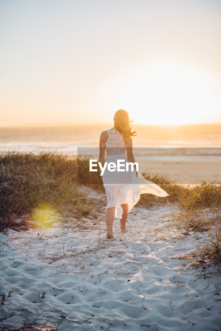 Rear view of young woman walking at beach against clear sky during sunset