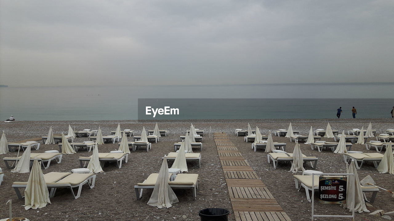 Deck chairs with parasols arranged at beach against cloudy sky
