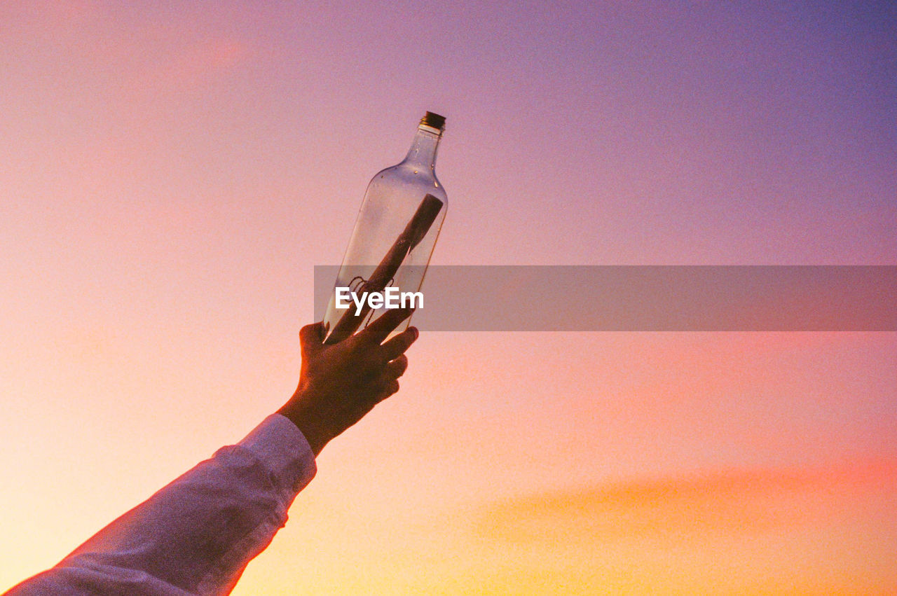 cropped hand of person holding bottle against sky during sunset