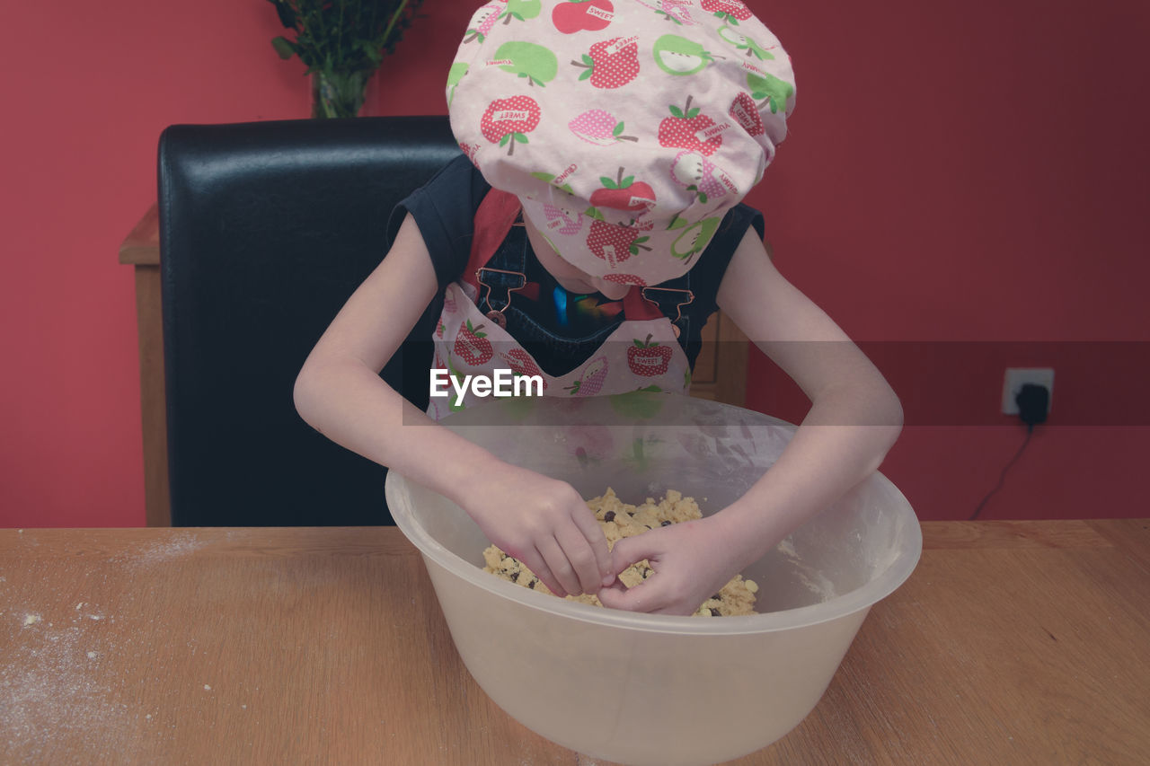 Girl wearing apron mixing batter in container on table at home