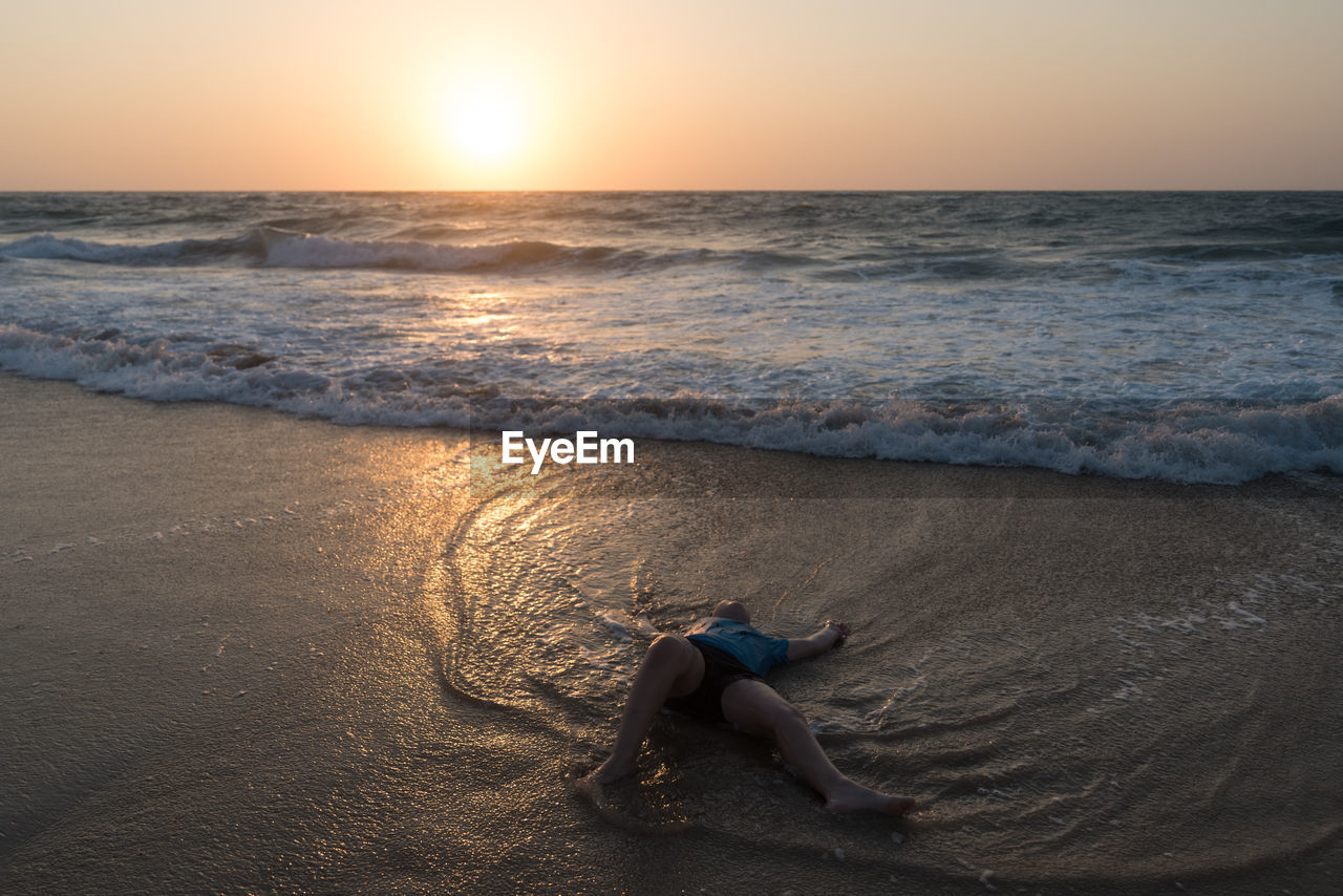 Boy lying down at beach against sky during sunset