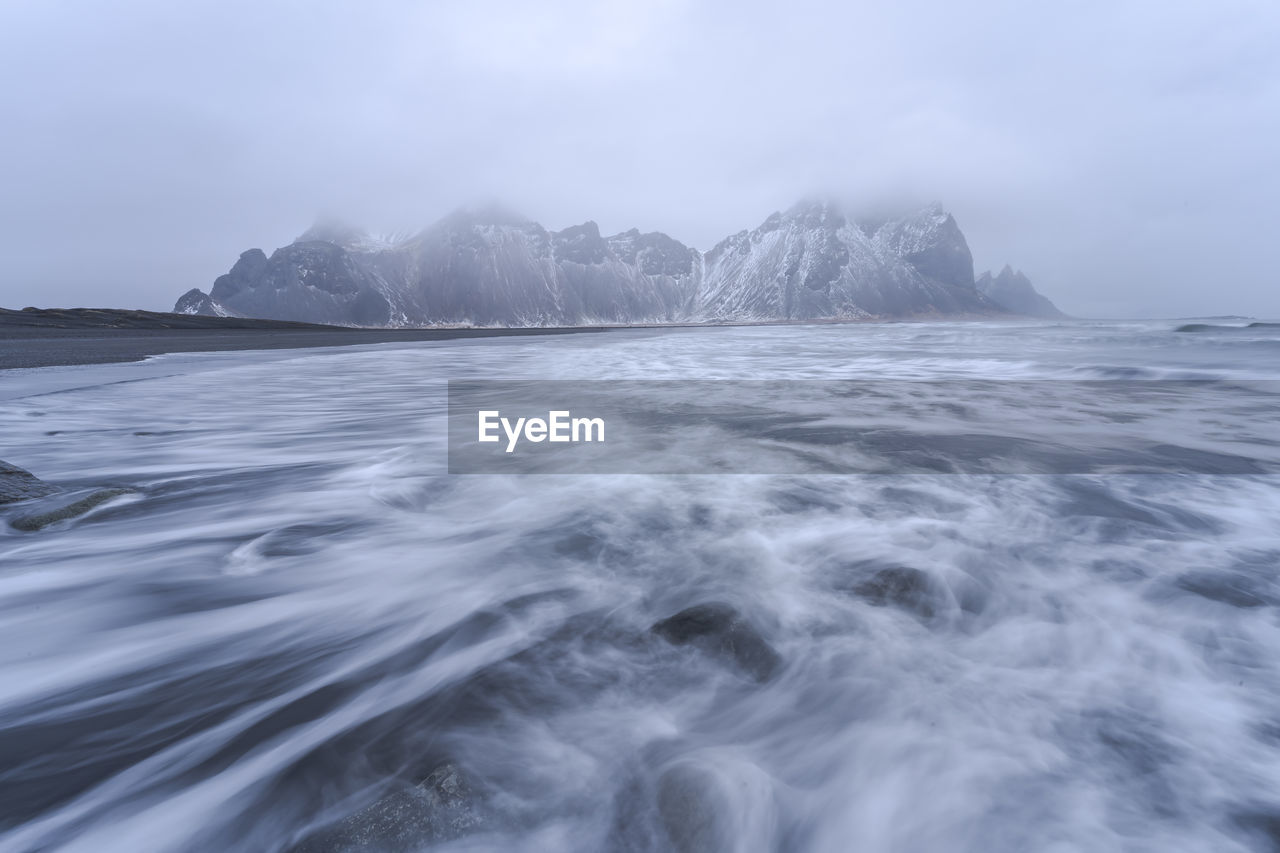 Picturesque scenery of powerful stormy sea water near rough rocky vestrahorn mountain formations on outcast misty day in stockness beach, iceland