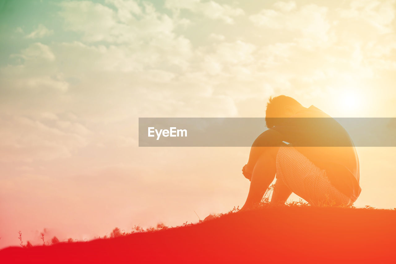 Side view of man sitting on land against orange sky during sunset