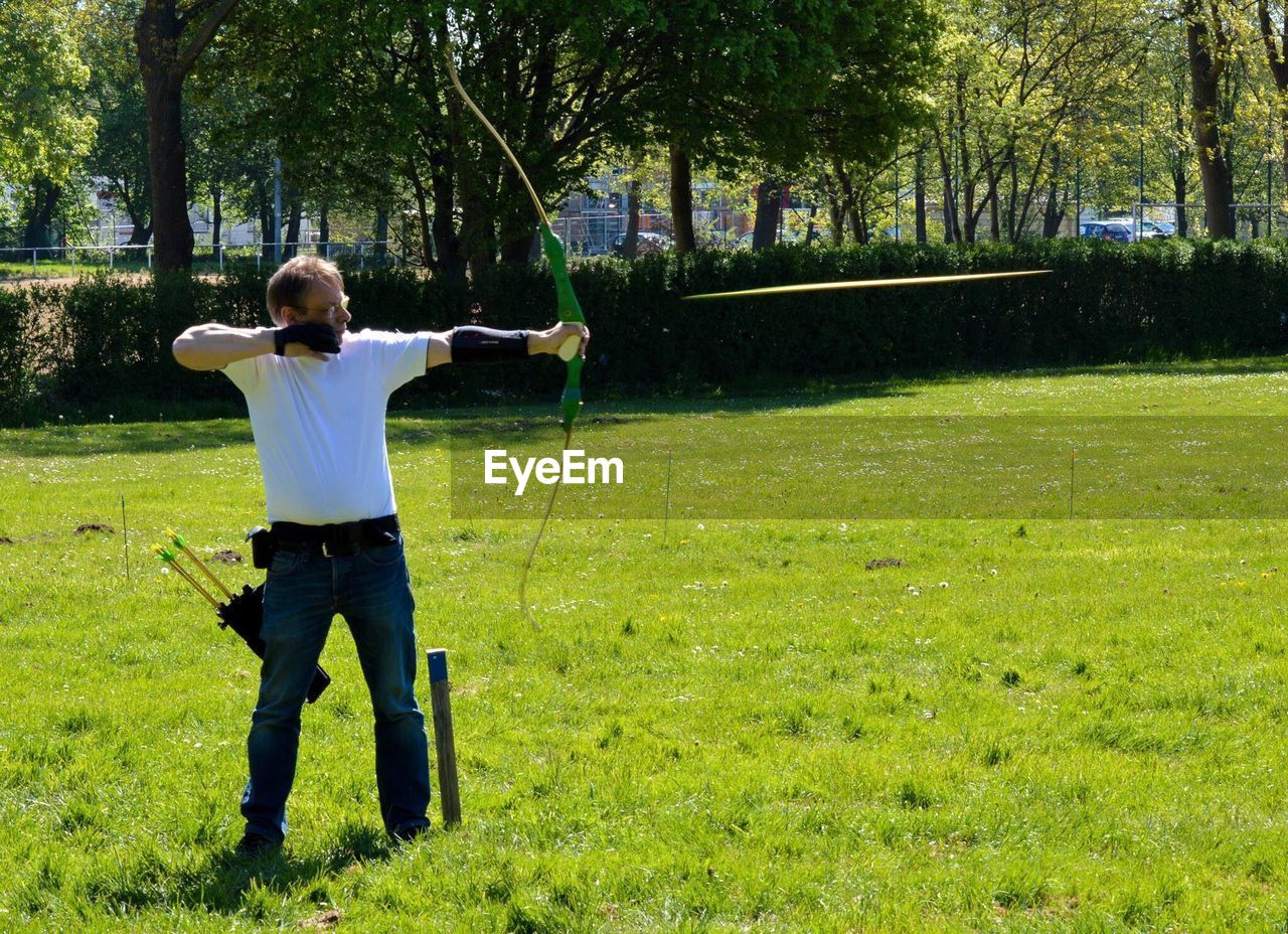 Full length of man shooting arrow on grassy field during sunny day