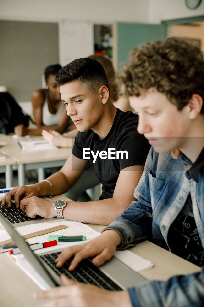 Male students using laptop while sitting by table in classroom