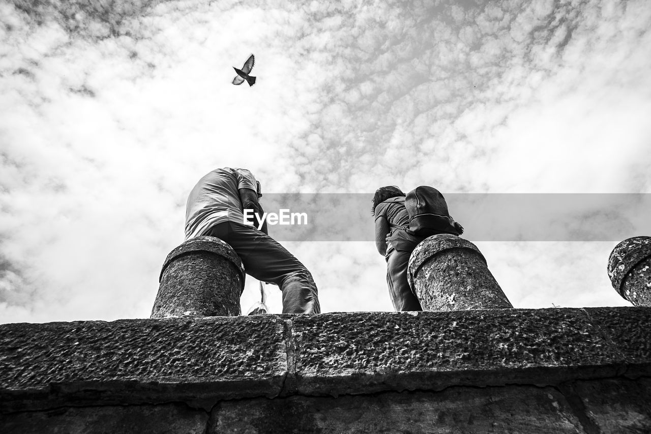 Low angle view of people sitting on bollard with bird flying against sky