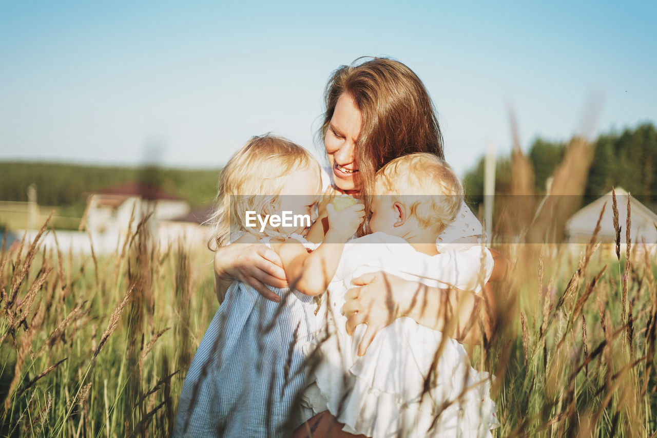 Young woman mom with baby girls twins on hands hug in backyard of cottage in sunset light