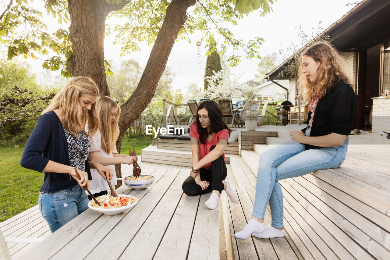 Teenage girls looking at friends mixing salad on wooden table in yard