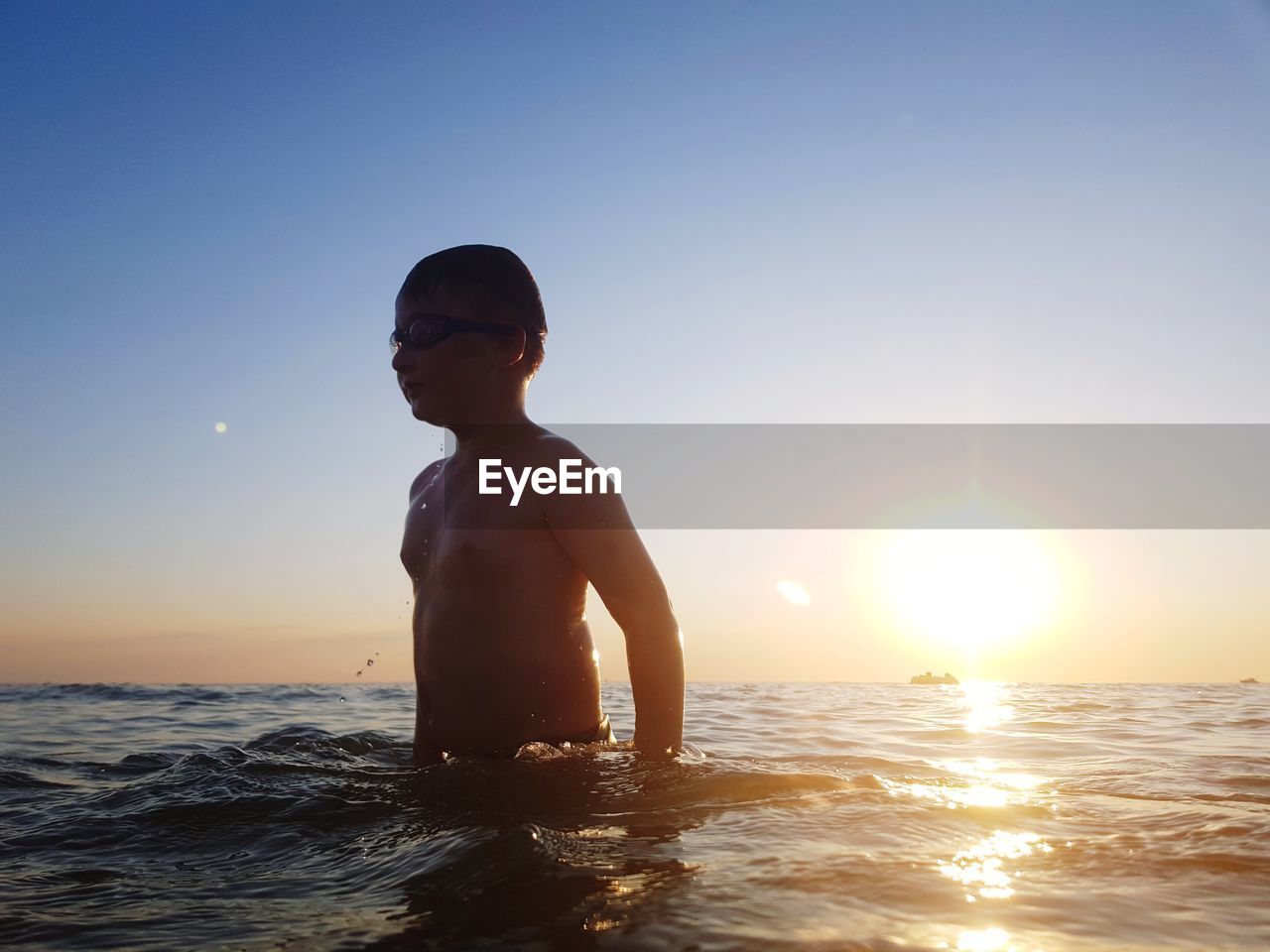 Shirtless boy standing in sea against clear sky during sunset