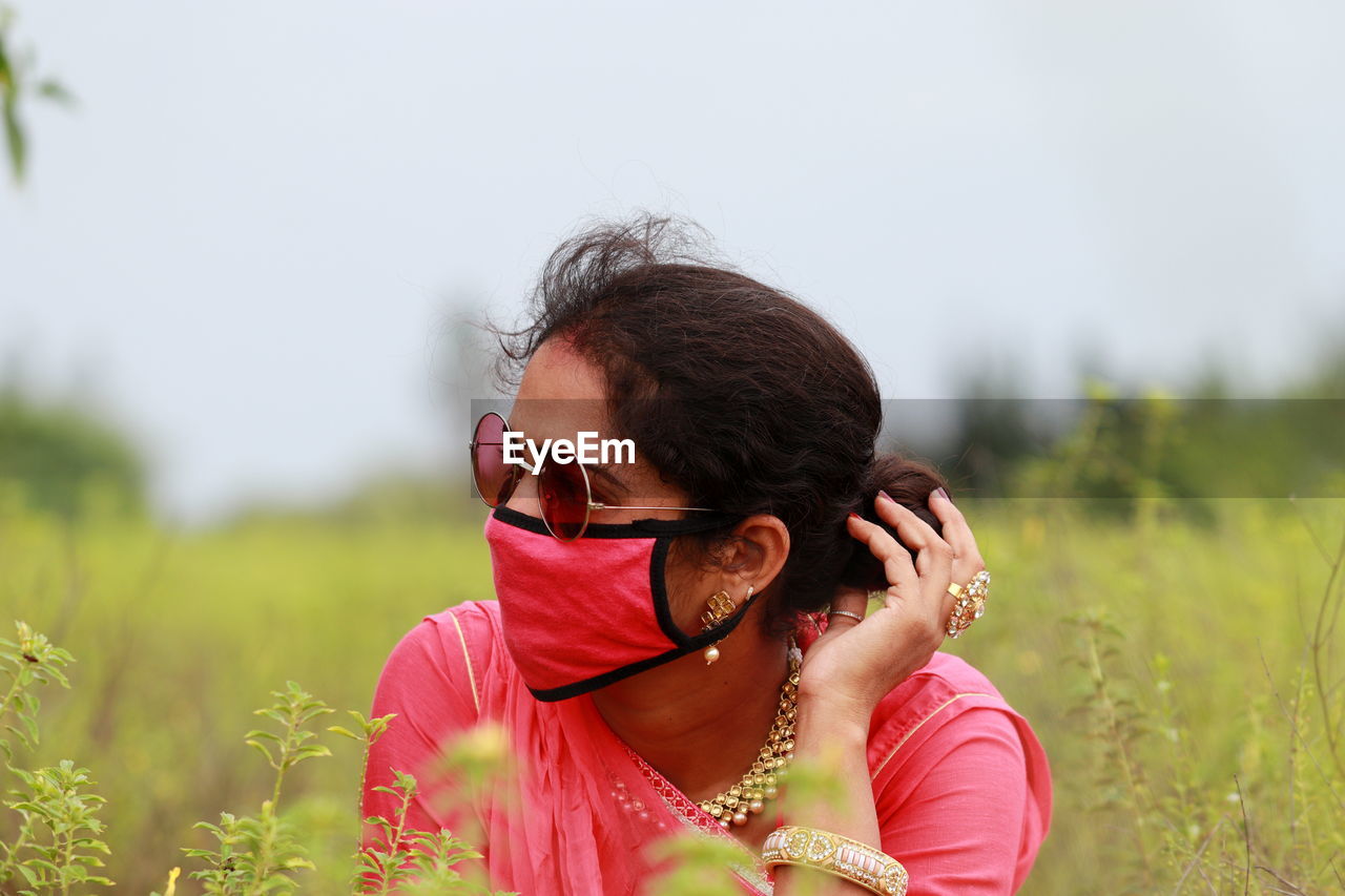 Young woman wearing mask looking away while sitting on field against sky