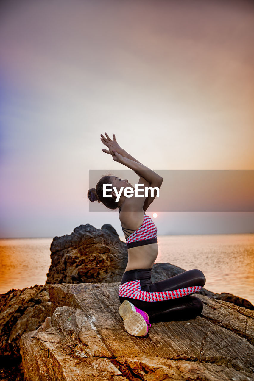 Woman exercising on rock against sea during sunset