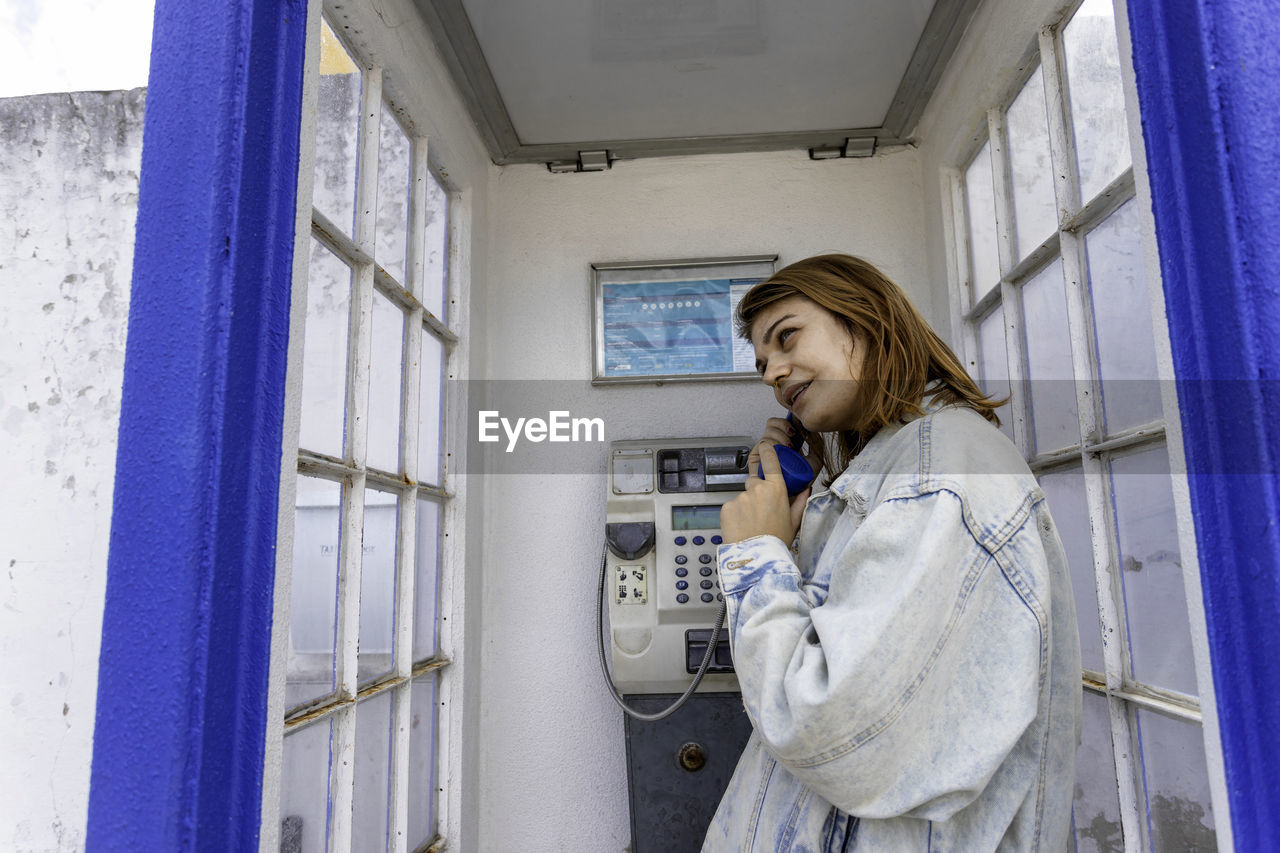 Smiling woman talking on telephone standing in telephone booth