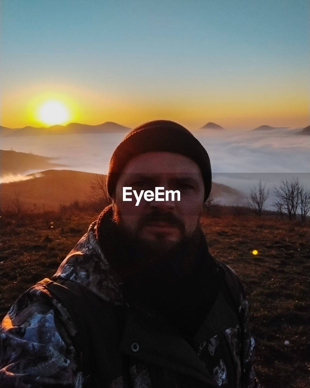 sky, one person, sunset, portrait, adult, nature, mountain, men, clothing, looking at camera, beauty in nature, leisure activity, scenics - nature, front view, landscape, sunlight, land, environment, sun, young adult, standing, facial hair, lifestyles, beard, non-urban scene, winter, outdoors, mountain range, orange color, evening, tranquility, hat, tranquil scene, jacket, travel, adventure, cold temperature, dusk, headshot, cloud, travel destinations, hiking, back lit, activity, copy space