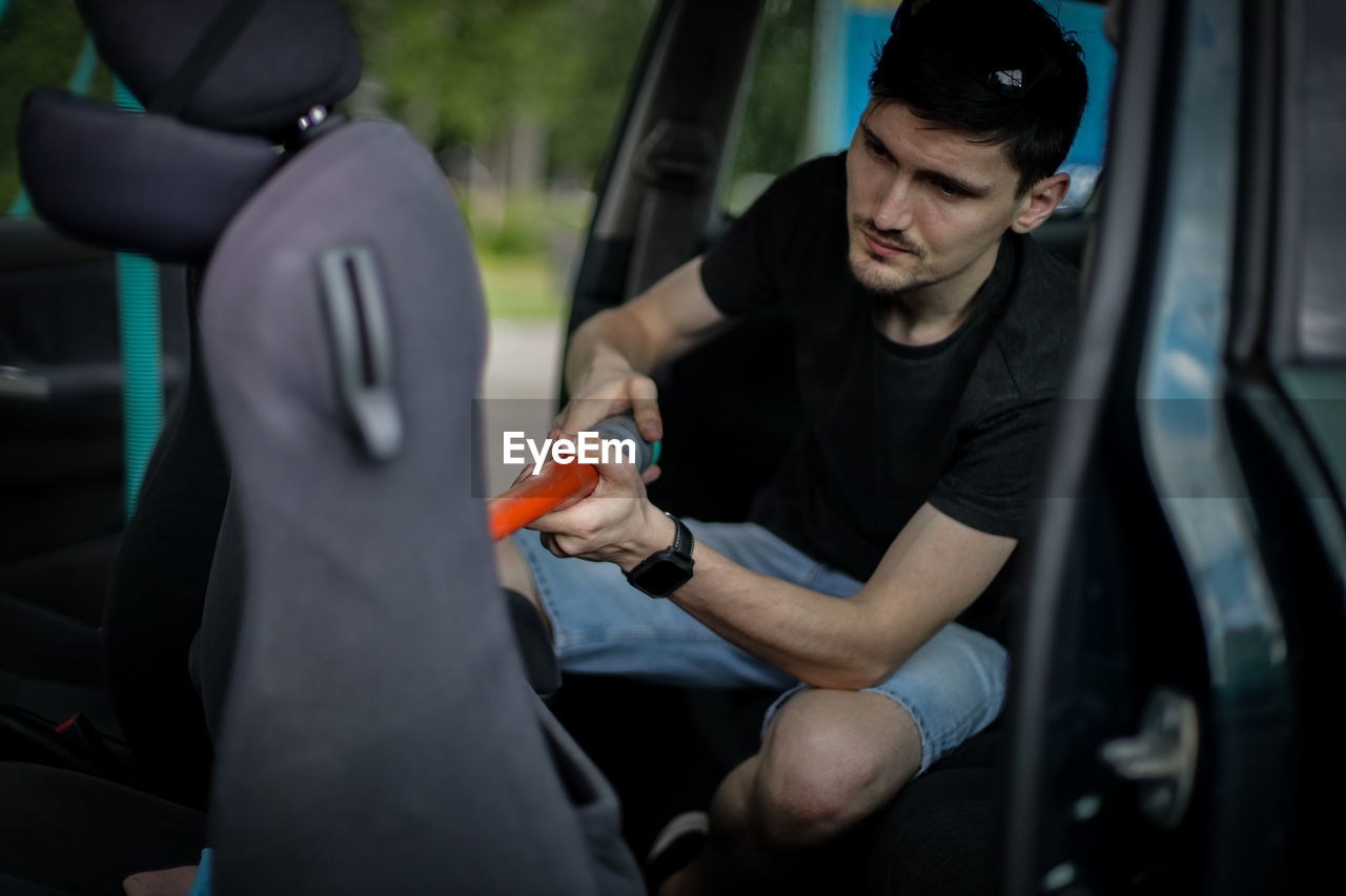 A young caucasian guy holds a tube with a nozzle in his hands and vacuums the seat in the car