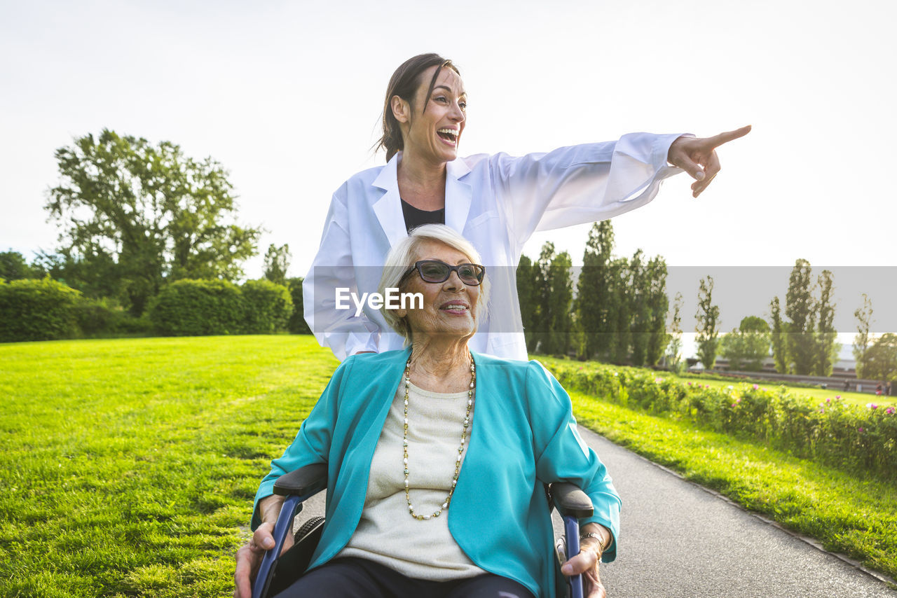 Cheerful female caretaker pointing while wheeling senior woman in park