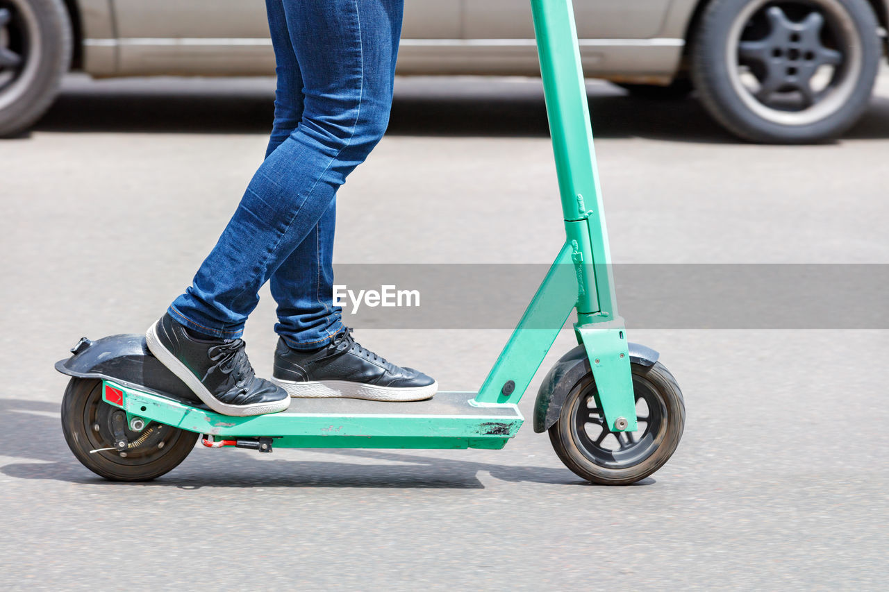 A man in blue jeans rides a rented electric scooter on a sunny day with a passing car .