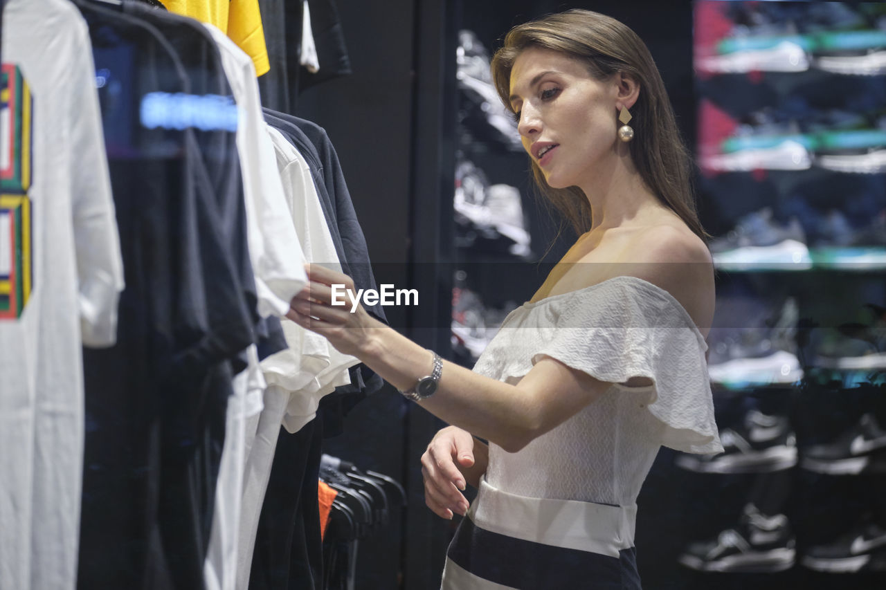 Side view of young stylish female standing near rack and choosing shirt while shopping in modern clothing store