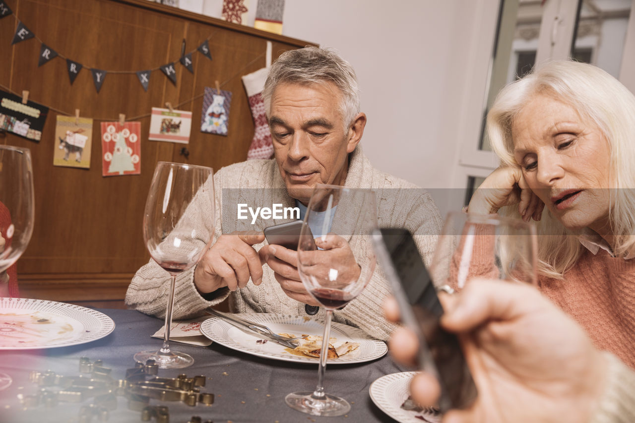 Annoyed senior woman with husband using smartphone after christmas dinner