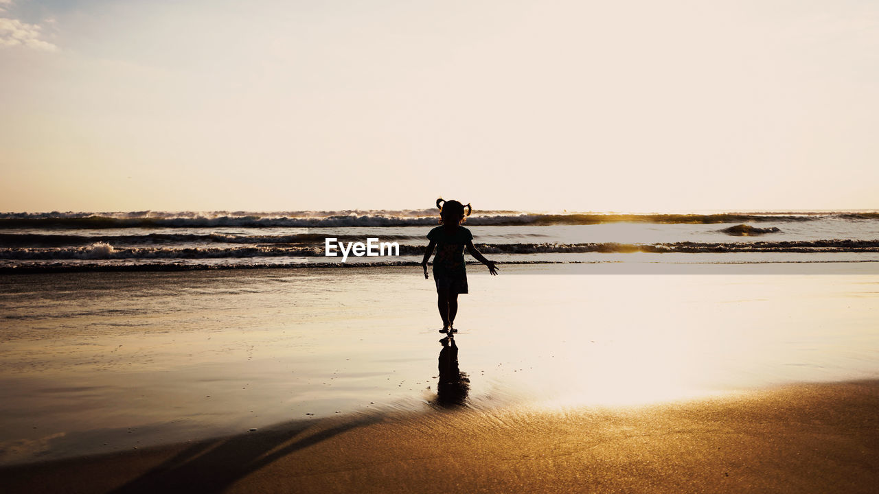 Child standing on beach against sky during sunset