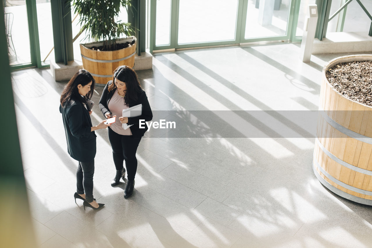 Female coworkers discussing over smart phones while standing at office corridor