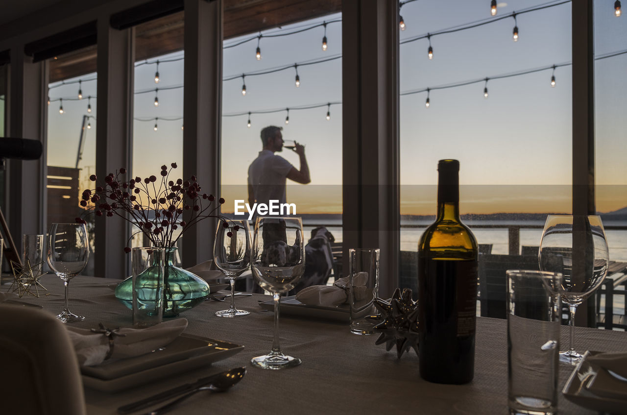 Man seen through window drinking wine while standing against sky during sunset