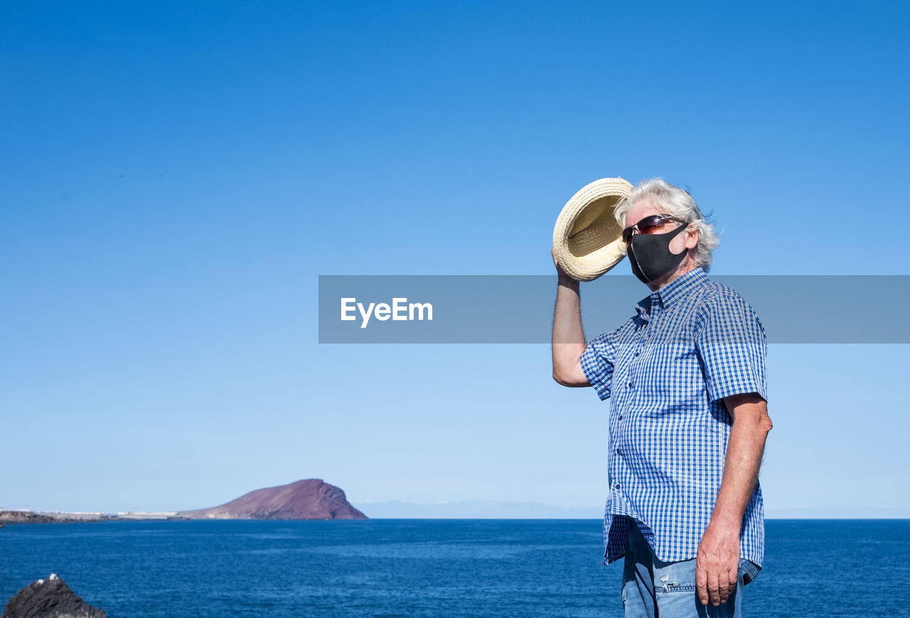 Man with face mask standing by sea against clear blue sky