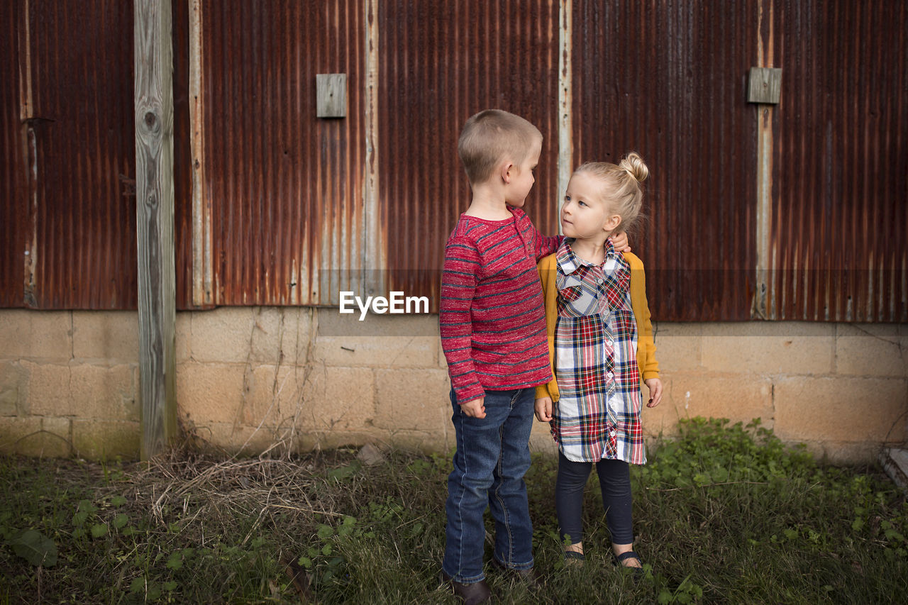 Siblings looking each other face to face while standing against abandoned building on field