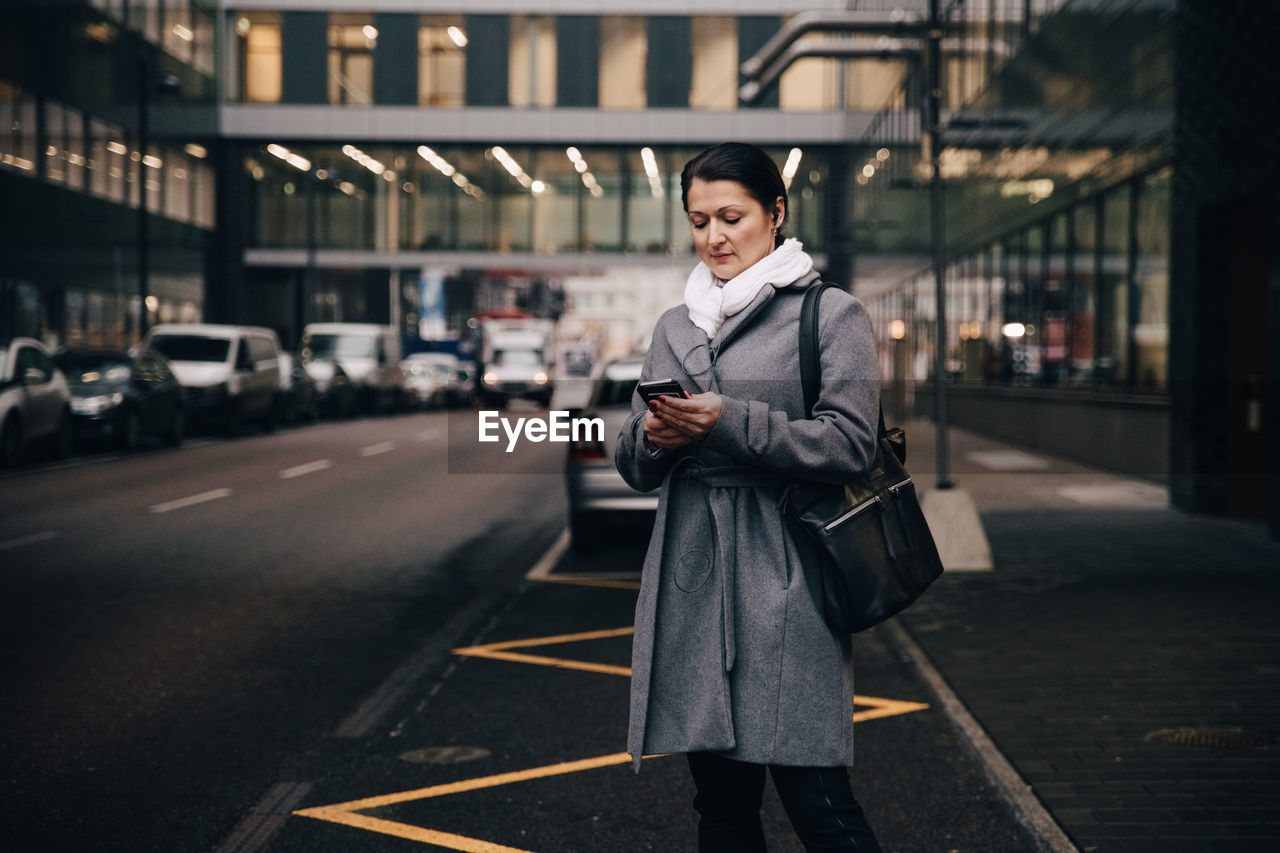 Businesswoman using smart phone while standing on city street against building