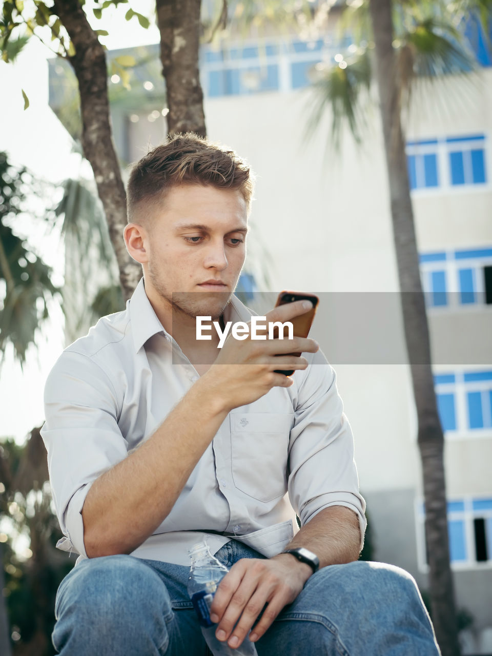 YOUNG MAN USING SMART PHONE WHILE SITTING ON TREE