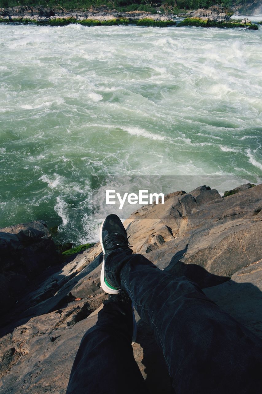 LOW SECTION OF MAN SITTING ON ROCK AT SEA SHORE