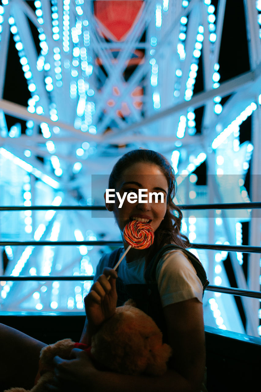 Pretty female teenager with teddy bear having fun in funfair and eating colorful lollipop while sitting in ferris wheel with blue illumination