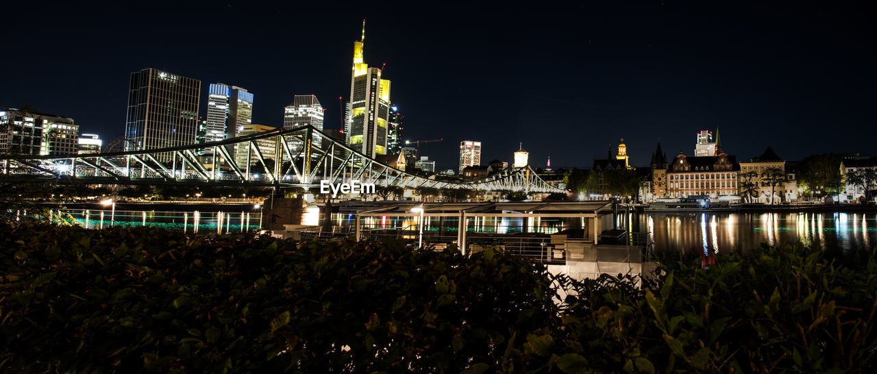 Illuminated modern buildings by river against sky at night