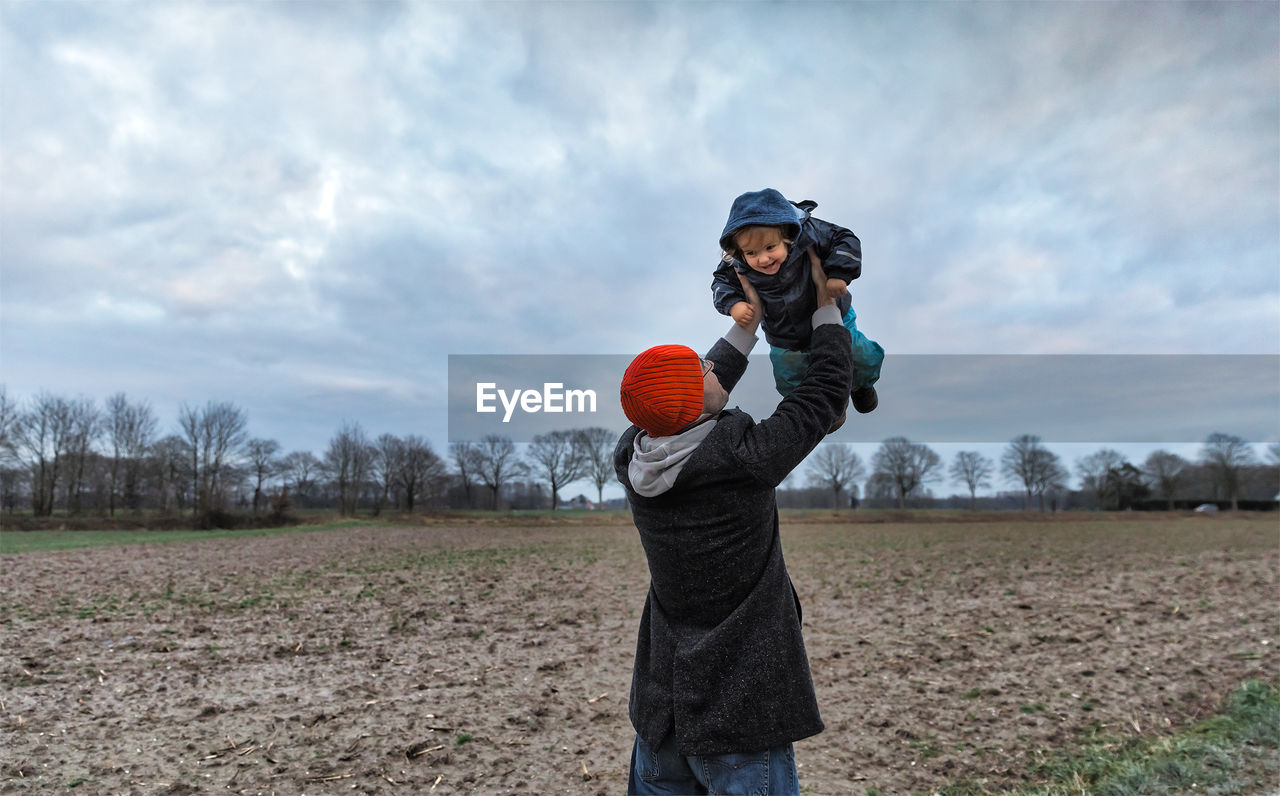 Father picking up son while standing on field against cloudy sky