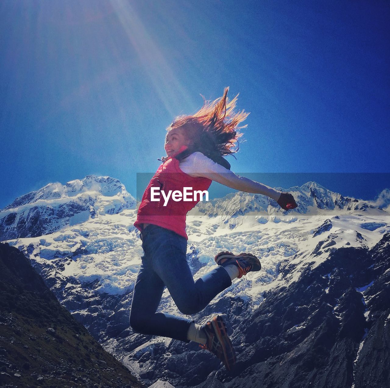 Low angle view of young woman jumping over snowcapped mountain against blue sky
