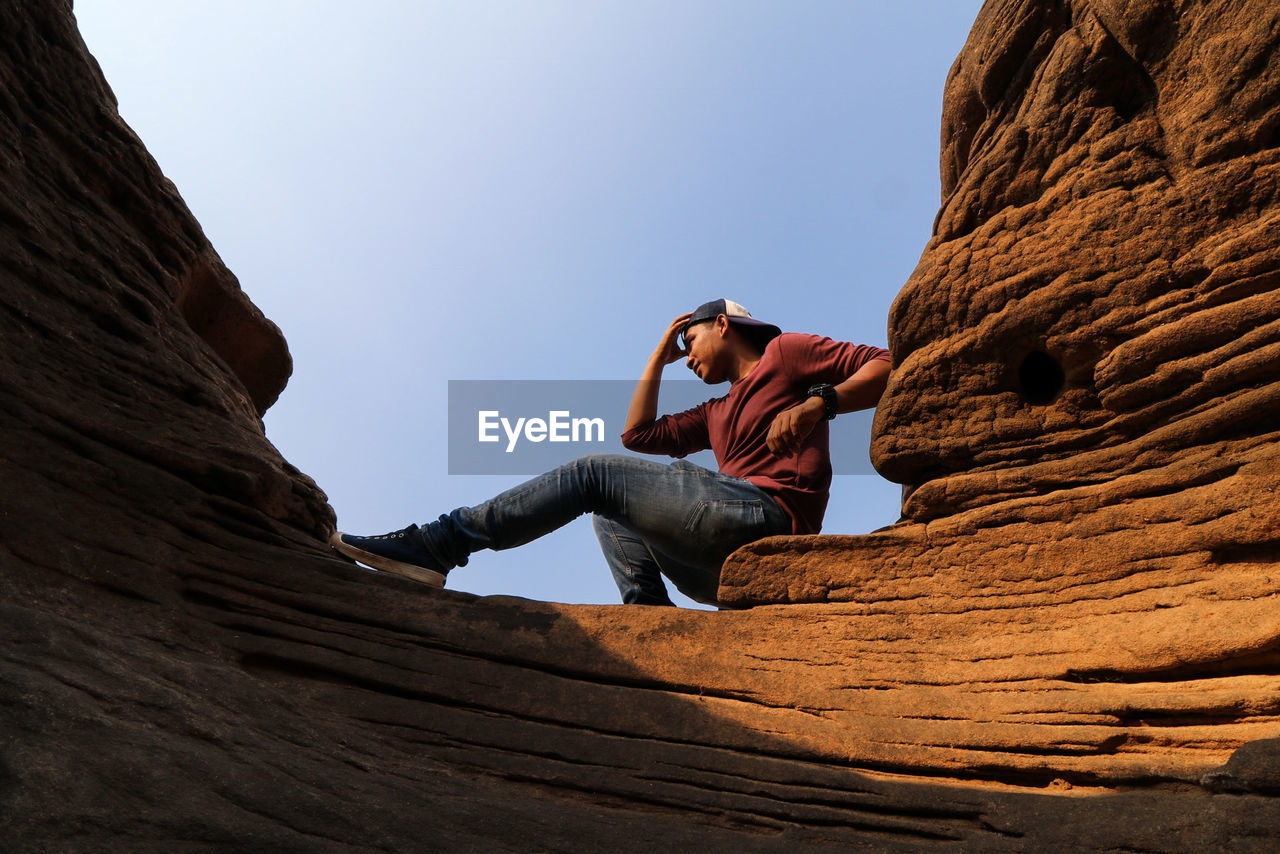 Low angle view of young man sitting on rock formation against sky