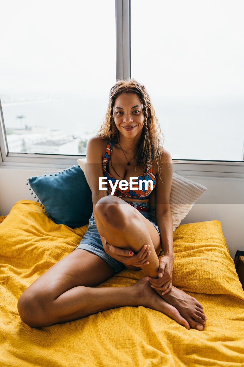 Young woman sitting on a bed in front of a window and smiling into the camera in a bedroom