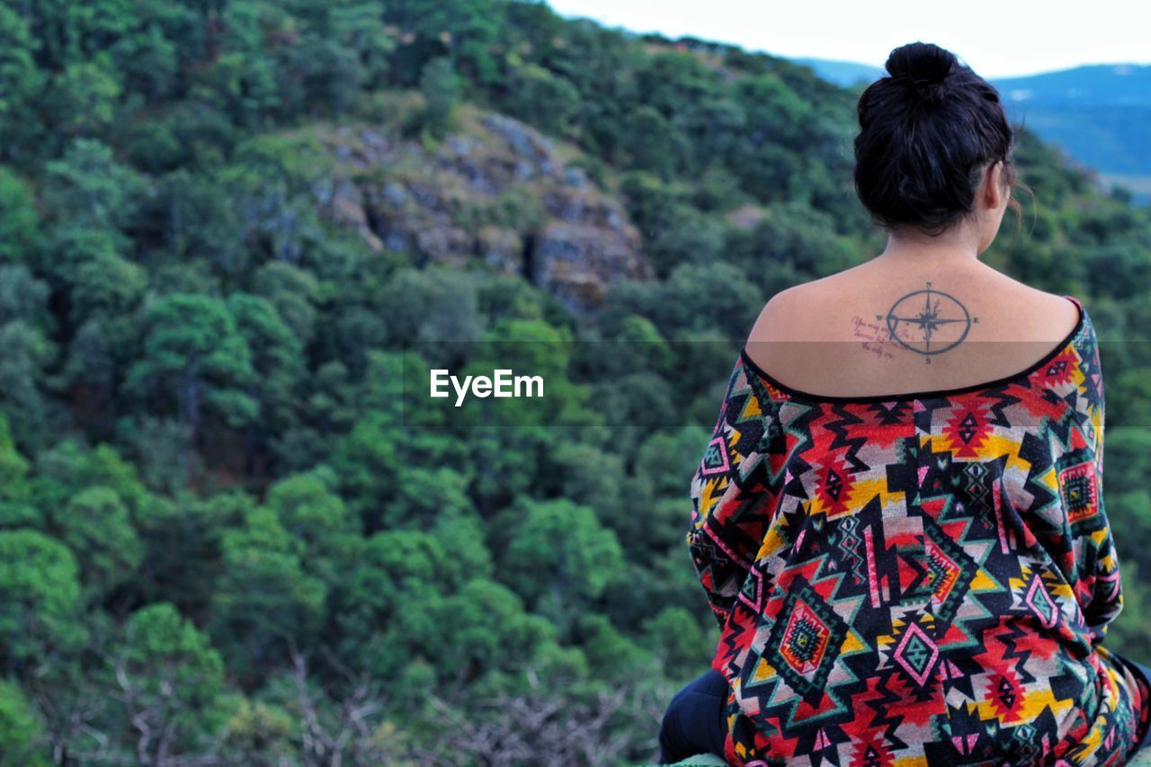 Rear view of young woman with tattoo on back sitting against trees