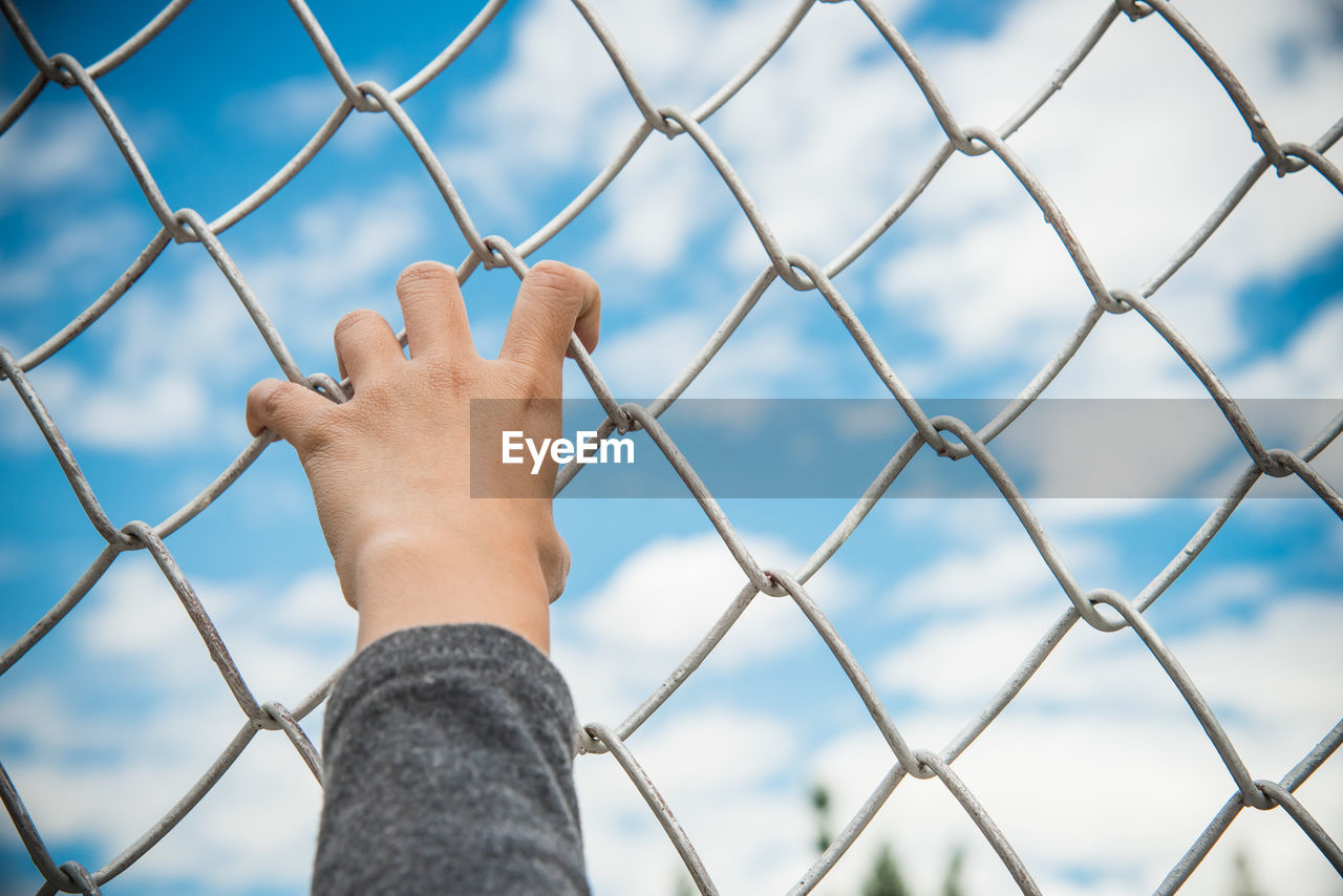 Cropped hand holding chainlink fence against sky