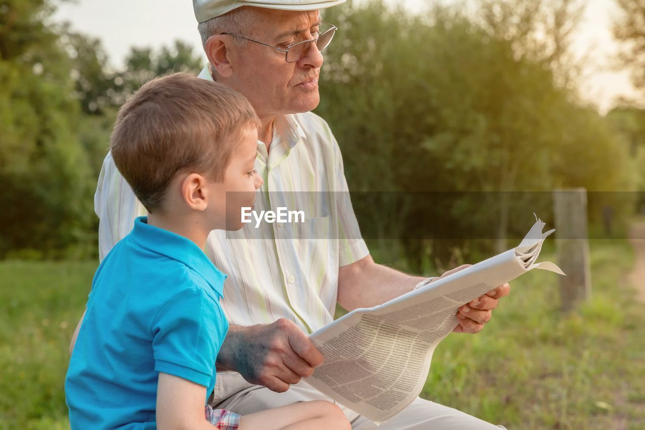 Grandfather reading newspaper while sitting by grandson in park