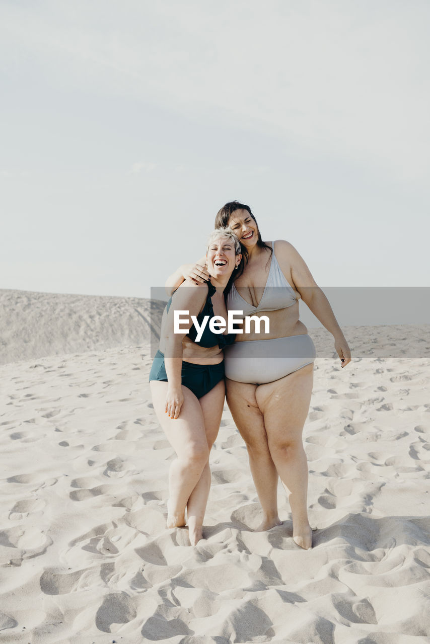 Glad curvy female friends in swimwear stand on sandy seashore and enjoying summer vacation together