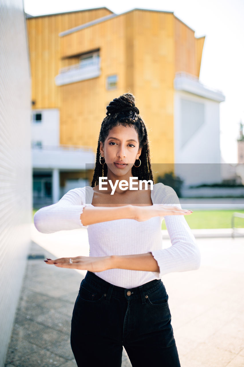 Portrait of young woman gesturing equal sign while standing against building